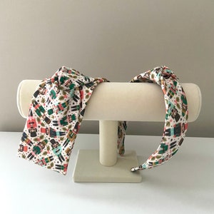 May include: Two white fabric headbands with a repeating pattern of colorful nutcracker figures on a white background. The headbands are displayed on a white headband stand.