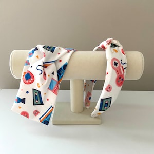 May include: Two white fabric headbands with a knot design. One headband has a retro video game and cassette tape print. The other headband has a retro video game controller and cassette tape print.