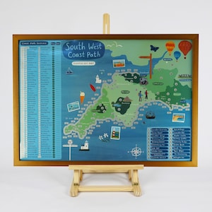 May include: A framed scratch-off map of the South West Coast Path, presented on a wooden easel. The map is a colorful illustration of the coastline, with landmarks, boats, and hot air balloons. The frame is a warm brown color.
