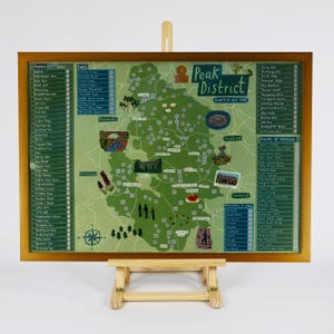 May include: A framed scratch-off map of the Peak District, presented on a small wooden easel. The map has a green background with illustrations of landmarks and points of interest, and lists of peaks and falls. The map is in a wooden frame.