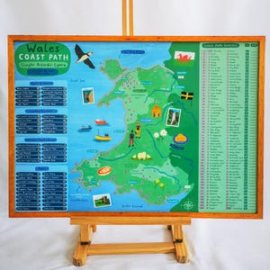 May include: A detailed map of the Wales Coast Path, displayed in a wooden frame. The map features illustrations of landmarks, boats, and wildlife, with text in English and Welsh. The artwork is supported by a wooden easel.