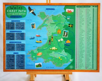 Op de afbeelding: Een ingelijste kaart van het Wales Coast Path, met illustraties van bezienswaardigheden, boten en dieren in het wild. De kaart bevat tekst in het Engels en Welsh, met een houten ezel die het kunstwerk ondersteunt. De lijst is bruin.