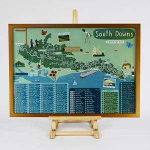 May include: A colourful illustrated scratch-off map of the South Downs National Park in England. The map features a green and blue colour scheme with a brown wooden frame. The map includes a list of peaks, forests, and points of interest. The text "South Downs Scratch Off Map" is printed at the top of the map.