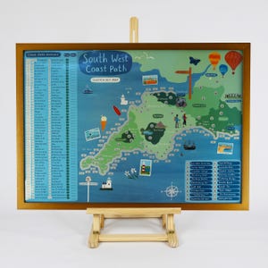 May include: A colourful scratch-off map of the South West Coast Path in England. The map features a blue ocean, green land, and various landmarks, including lighthouses, boats, and a hot air balloon. The map is framed in gold and displayed on a wooden easel.