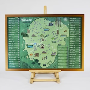 May include: A framed map of Dartmoor, with a green illustrated landscape and place names. The map is displayed on a wooden easel, showcasing a detailed depiction of the region. The map includes illustrations of landmarks.