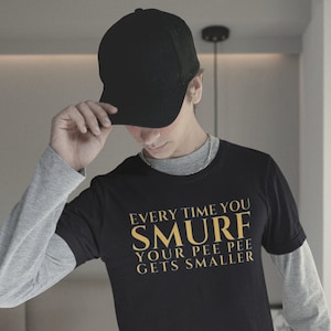 May include: Black t-shirt with gold text that reads "Every time you SMURF your pee pee gets smaller." The shirt is worn over a gray long-sleeved shirt. The person is wearing a black baseball cap and gray pants.