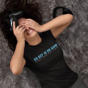 May include: Black t-shirt with the text "GG REP JG NO GANK noob team bot sleep" in light blue. The person is holding a black game controller and wearing gray pants. A pair of black headphones is also visible.