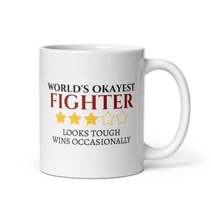 May include: White ceramic mug with a large handle. The mug features the text "WORLD'S OKAYEST FIGHTER" in black and red, with four gold stars. Below the text is "LOOKS TOUGH WINS OCCASIONALLY".