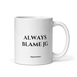May include: White ceramic mug with the text "ALWAYS BLAME JG" in black, with the hashtag "#gamerlaws" below. The mug has a curved handle and a glossy finish, suitable for hot or cold beverages.
