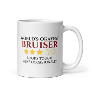May include: White ceramic mug with the text "WORLD'S OKAYEST BRUISER" in black and red, with gold star graphics. The mug also has the text "LOOKS TOUGH WINS OCCASIONALLY". The mug has a white handle.