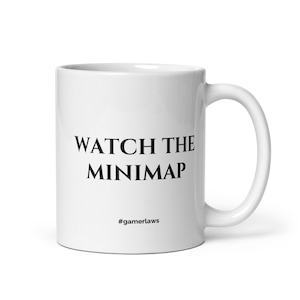 May include: White ceramic mug with the text "WATCH THE MINIMAP" in black, along with the hashtag "#gamerlaws". The mug has a curved handle and is designed for hot beverages. A perfect gift for gamers.
