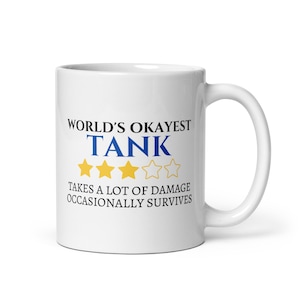 May include: A white ceramic mug with a curved handle. The mug features the text "WORLD'S OKAYEST TANK" in black and blue, with three yellow stars. Below the text is the phrase "TAKES A LOT OF DAMAGE OCCASIONALLY SURVIVES."