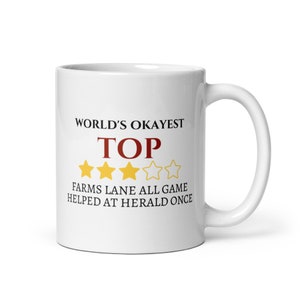 May include: A white ceramic mug with a curved handle. The mug features the text "WORLD'S OKAYEST TOP" in black and red, with four gold stars. Below the text is "FARMS LANE ALL GAME HELPED AT HERALD ONCE."