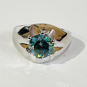 May include: A silver ring with a large, round, teal-colored gemstone. The ring has a wide band and a polished finish, reflecting light. The gemstone is faceted, creating a sparkling effect. The ring is set on a white surface.