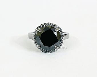 May include: A black gemstone ring with a halo of smaller stones. The large, round, black center stone is surrounded by a ring of smaller, clear stones. The band is a silver color, and the ring is set against a white background.