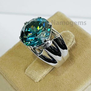May include: A large, silver ring featuring a vibrant, teal-colored gemstone. The gemstone is oval-shaped and set in a four-prong setting. The ring's band is thick and polished, with a modern design. The text "Namogems" is visible on the right side of the ring.