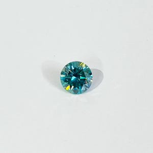 May include: A round, faceted gemstone with a vibrant teal color. The gem has a brilliant cut, reflecting light to create a sparkling effect. The stone is set against a plain white background, highlighting its color and clarity.