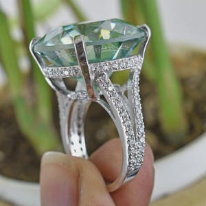 May include: A silver ring with a large, light green gemstone in a cushion cut. The ring is embellished with small, clear stones along the band and around the main stone. The ring is held up against a blurred green background.