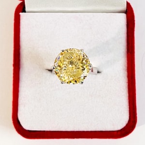 May include: A silver ring with a large, round, yellow gemstone. The ring is set in a six-prong setting and is displayed in a red velvet ring box with a white interior.