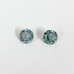 May include: Two round, teal-colored gemstones with multiple facets are displayed against a white background. The gems have a brilliant cut, reflecting light and creating a sparkling effect. The stones are likely for jewelry.