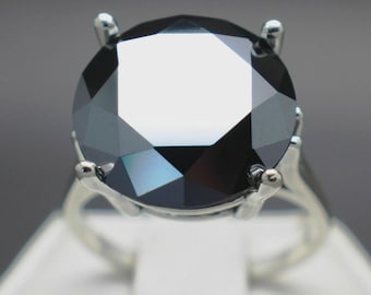 May include: A close-up of a silver ring featuring a large, faceted black gemstone. The gemstone is round with multiple reflective surfaces. The ring's band is a simple, polished silver design, with four prongs securing the stone.
