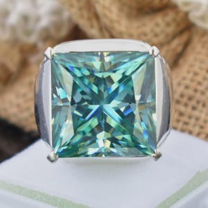 May include: A square-cut, teal-colored gemstone ring with a silver band. The gemstone has multiple facets that reflect light, creating a sparkling effect. The ring is set on a white surface.