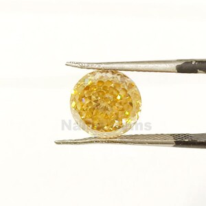 May include: A round, faceted yellow gemstone is held by a pair of tweezers against a white background. The gemstone has a brilliant cut, reflecting light and showcasing its golden color. The tweezers are silver.