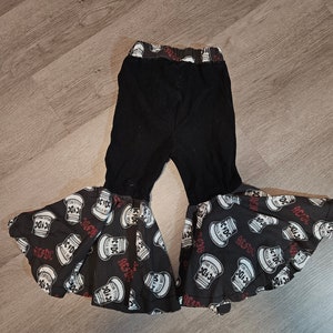 Puede incluir: Pantalón negro de campana con una banda roja y blanca alrededor de la cintura. El pantalón tiene un estampado en blanco y negro de una lata con el texto "AC/DC" y "For Those About To Rock We Salute You".
