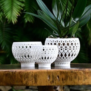 Puede incluir: Tres macetas de cerámica blanca con intrincados diseños geométricos. Las macetas están dispuestas sobre una mesa de madera con una planta verde de hojas en la maceta más grande.