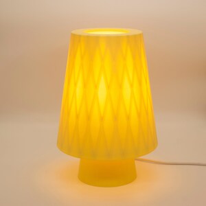 May include: A yellow table lamp with a geometric patterned shade. The lamp has a wide base and a white cord.