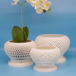 Op de afbeelding: Drie witte keramische plantenpotten met een tralieontwerp. De potten zijn in elkaar genest, met de grootste pot onderaan en de kleinste pot bovenaan. Een witte orchideeënplant bevindt zich in de grootste pot.