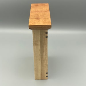 May include: Side view of a wooden box with a light wood finish. The box has a simple design with a flat top and a rectangular shape. The box is made of two different types of wood, one light and one darker. The box has a small black detail on the side.