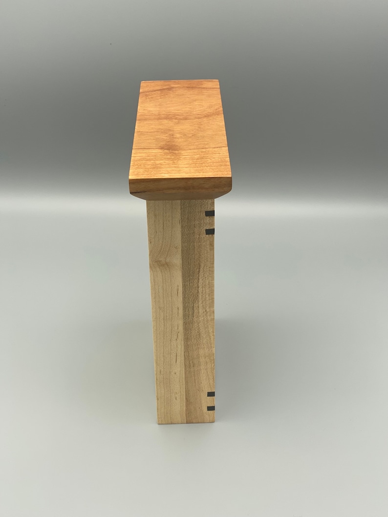 May include: Side view of a wooden box with a light wood finish. The box has a simple design with a flat top and a rectangular shape. The box is made of two different types of wood, one light and one darker. The box has a small black detail on the side.
