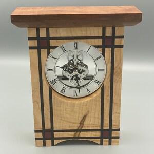 May include: A wooden clock with a silver face and Roman numeral markings. The clock is framed with a geometric design of black and brown wood with red accents.
