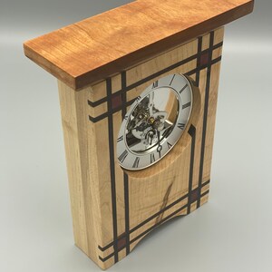 May include: A wooden clock with a white face and black Roman numeral markings. The clock is encased in a wooden frame with a geometric design featuring black and red accents.