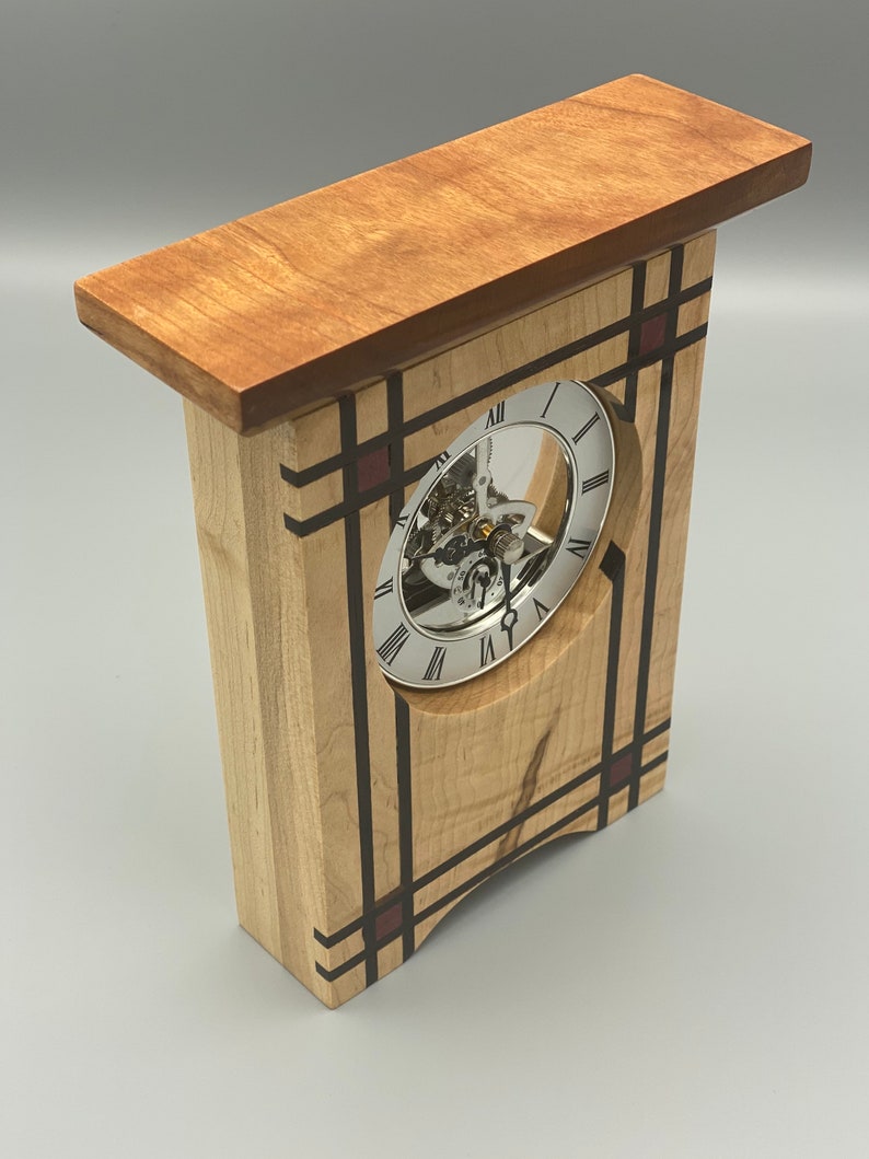 May include: A wooden clock with a white face and black Roman numeral markings. The clock is encased in a wooden frame with a geometric design featuring black and red accents.