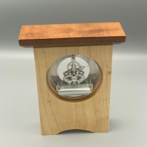 May include: A wooden clock with a clear face and visible gears. The clock is mounted on a wooden stand with a cherry wood top.