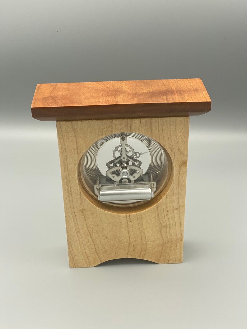 May include: A wooden clock with a clear face and visible gears. The clock is mounted on a wooden stand with a cherry wood top.