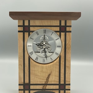 May include: A wooden clock with a silver face and Roman numeral markings. The clock is made of maple and walnut wood with black and red accents.