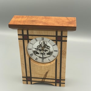 May include: A wooden clock with a silver face and Roman numeral markings. The clock is set in a wooden frame with a geometric design featuring black and red accents.