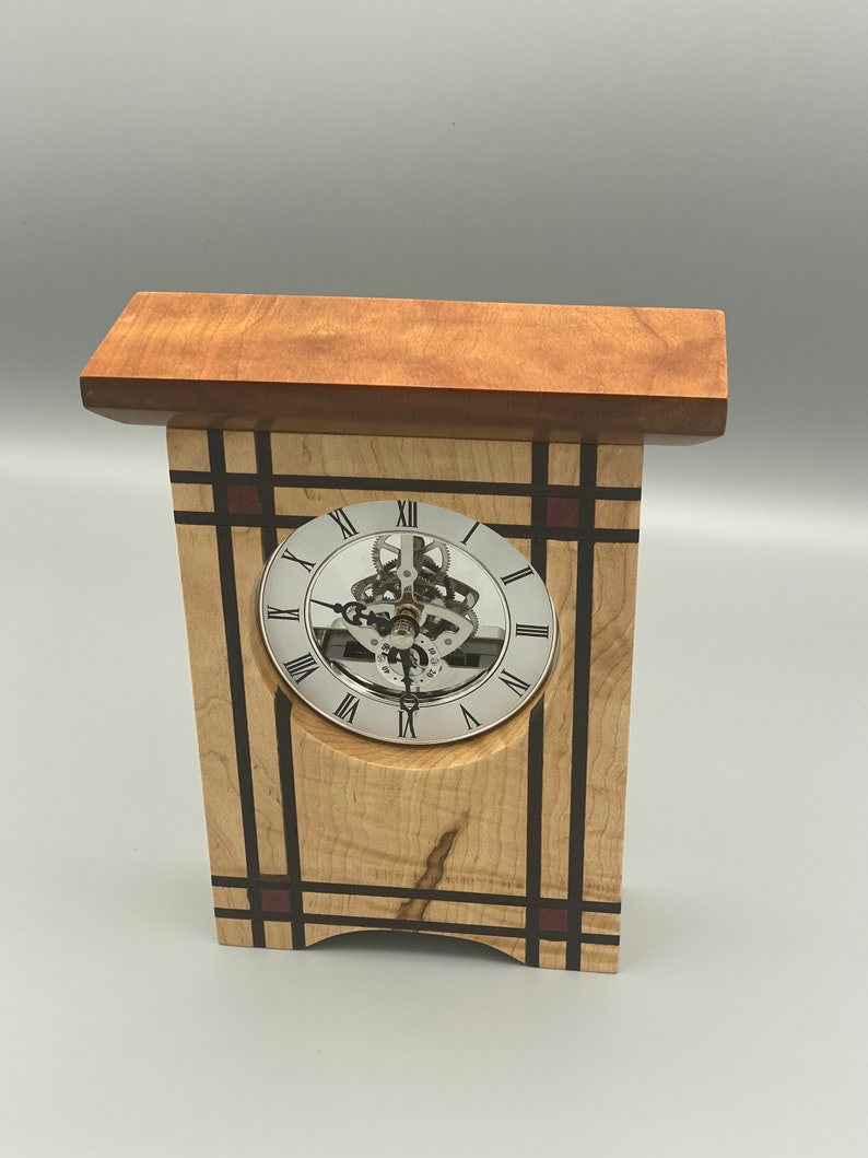 May include: A wooden clock with a silver face and Roman numeral markings. The clock is set in a wooden frame with a geometric design featuring black and red accents.