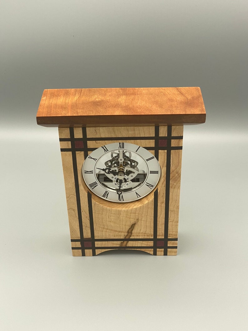 May include: A wooden clock with a white face and black Roman numeral markings. The clock is set in a wooden frame with a geometric pattern of black and red stripes. The clock is sitting on a gray surface.