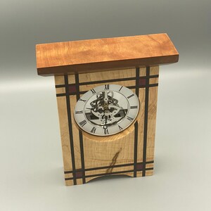 May include: A wooden clock with a Roman numeral clock face and a visible gear mechanism. The clock is made of maple wood with red and black accents.