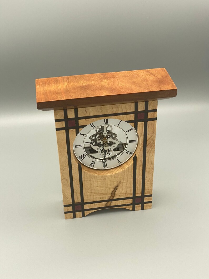 May include: A wooden clock with a Roman numeral clock face and a visible gear mechanism. The clock is made of maple wood with red and black accents.