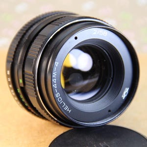 Pode incluir: Uma lente Helios-44M-4 58mm f/2 preta com um anel e marcas prateadas. A lente tem um anel de abertura preto com marcas brancas.