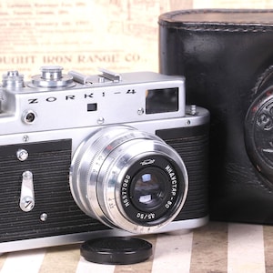 May include: A vintage Zorki-4 camera with a silver body and black accents, featuring a prominent lens and a black lens cap. A black leather case is visible on the right. The camera is a classic piece of photography equipment.
