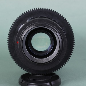 Puede incluir: Primer plano de una lente negra con un anillo exterior en forma de engranaje y un centro de cristal transparente. Un peque&ntilde;o punto rojo es visible en el lateral. La lente est&aacute; sobre un fondo verde claro.