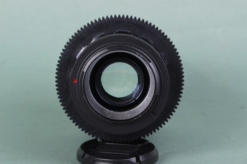 Puede incluir: Primer plano de una lente negra con un anillo exterior en forma de engranaje y un centro de cristal transparente. Un peque&ntilde;o punto rojo es visible en el lateral. La lente est&aacute; sobre un fondo verde claro.