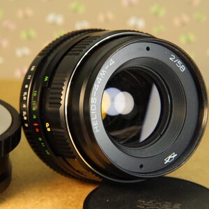 Pode incluir: Uma lente de c&acirc;mera preta com um anel prateado e o texto "HELIOS-44M-4 2/58" impresso na lateral.