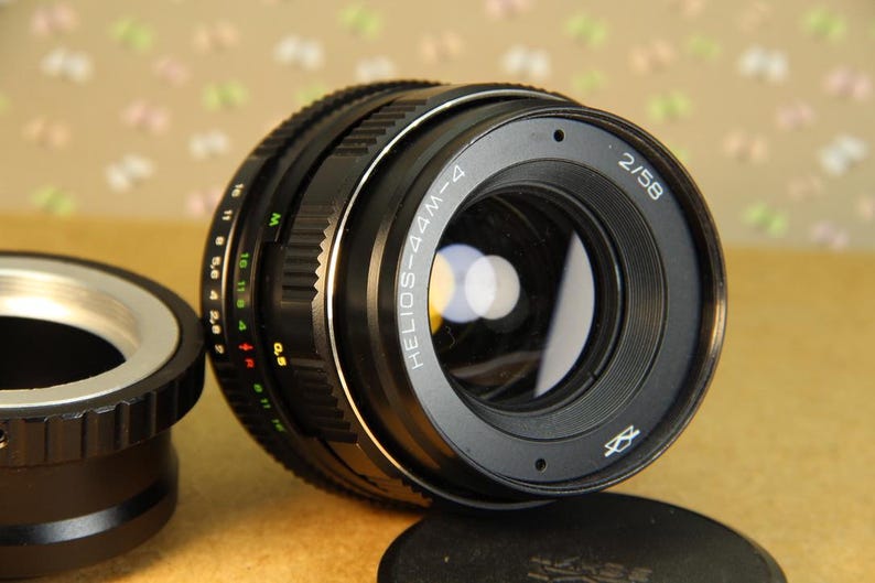 Pode incluir: Uma lente de c&acirc;mera preta com um anel prateado e o texto "HELIOS-44M-4 2/58" impresso na lateral.